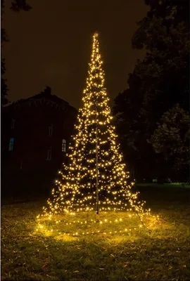 A tall, cone-shaped Christmas tree made of lights stands in a grassy area at night, with a building in the background.