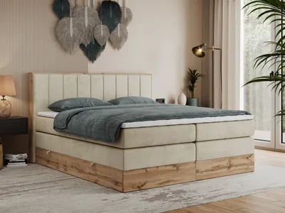 A bedroom scene featuring a king-size bed with a beige upholstered headboard and base, complemented by wood-tone accents. The bed is dressed with gray bedding and pillows. Decorative feather wall art hangs above the headboard, and a floor lamp and a potted