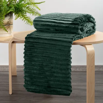 A dark green ribbed blanket is folded and draped over a light wood side table, with a potted fern in the background.