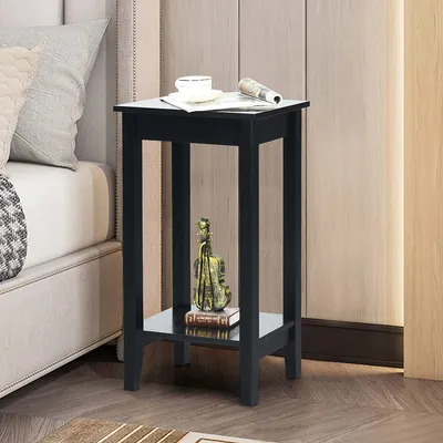 A black side table with a shelf is placed next to a bed, with a cup and newspaper on top and a decorative item on the shelf.