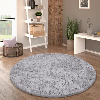 A round, gray, shaggy rug is placed on a wooden floor in a living room setting. A ladder shelf with decorative items, a basket with a pillow, and a plant are visible in the background.