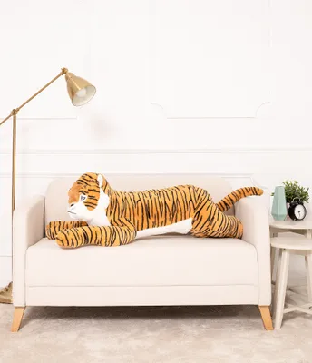 A large plush tiger toy is lying on a sofa in a room with a floor lamp and a side table.