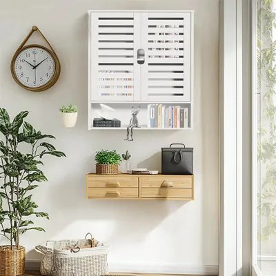 A white wall-mounted cabinet with slatted doors, a small shelf, and two drawers below, decorated with plants, books, and a clock.