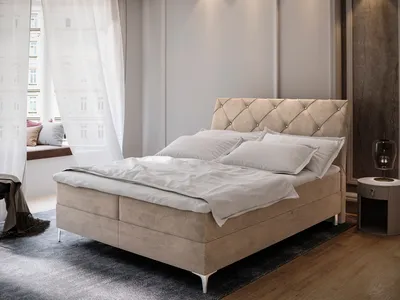 A bedroom scene featuring a beige boxspring bed with white bedding, a window with sheer curtains, and a bedside table with a lamp.