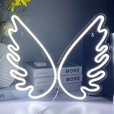 A white neon sign shaped like angel wings is displayed on a shelf with books and a vase of yellow flowers.