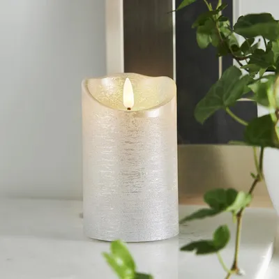 A close-up of a silver LED candle with a flickering flame, placed on a white surface with a plant in the foreground and a blurred background.
