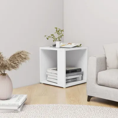 A white cube-shaped side table is placed in a corner of a room, next to a sofa. Books are stacked inside the table, and a plant and magazine are on top.