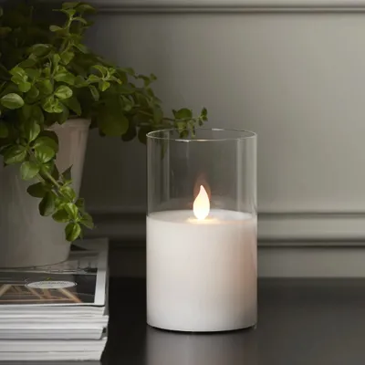 A white LED candle flickers inside a glass cylinder, placed on a dark surface next to a stack of magazines and a potted plant.