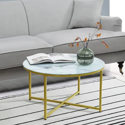 A round coffee table with a marble-like top and gold legs is placed on a striped rug in front of a gray sofa. A book and a vase with branches are on the table.