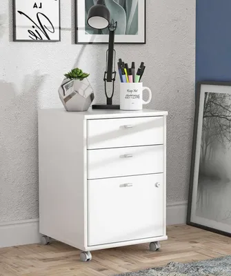 A white filing cabinet with three drawers and a cabinet door, placed on a wooden floor next to a wall with framed pictures and a desk lamp.