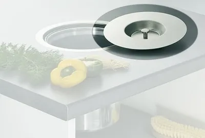 A close-up view of a sink drain cover on a countertop, with some vegetables in the background.