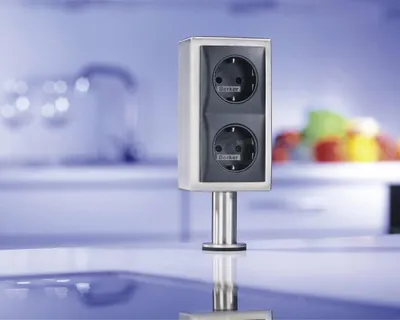 A stainless steel power outlet with two sockets, mounted on a countertop in a modern kitchen.