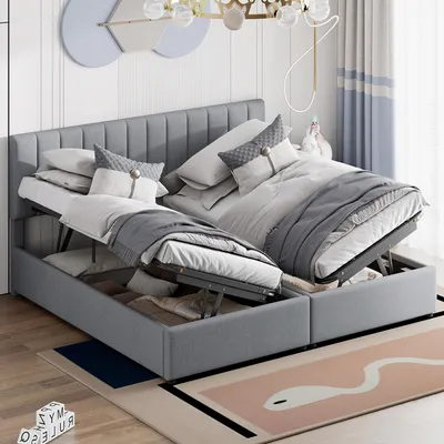 A bedroom setting with a grey upholstered bed frame, featuring two lift-up storage compartments. The bed is made with white and grey bedding and pillows. A decorative wall art and curtains are visible in the background.