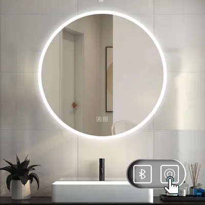 A round LED mirror with integrated lighting is mounted on a tiled wall above a white sink. The mirror reflects a doorway and a framed picture. A small plant is on the left, and a bottle of diffuser is on the right.