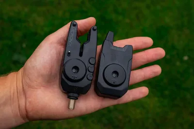 A hand holding two black fishing bite alarms outdoors.