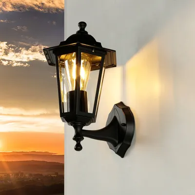 A black outdoor wall lamp is mounted on a white wall, with a sunset sky in the background.