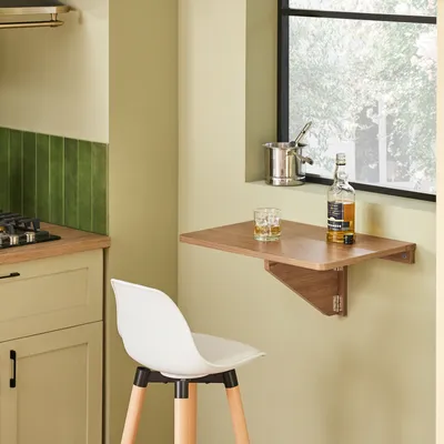 A small folding table mounted on a wall next to a window, with a bottle of whiskey and a glass on it. A white bar stool is in the foreground.