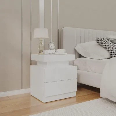 A white nightstand with two drawers is placed next to a bed in a bedroom. The nightstand has a lamp, a clock, and some books on top.