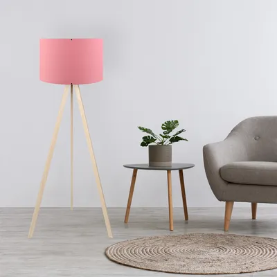 A pink floor lamp with a wooden tripod base stands next to a small side table with a potted plant and a gray armchair in a room.
