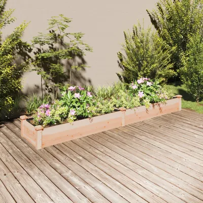 A wooden planter box filled with plants and flowers sits on a wooden deck, with a wall and bushes in the background.