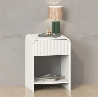 A white nightstand with a drawer and an open shelf, placed against a textured gray wall. A lamp and books are on top.