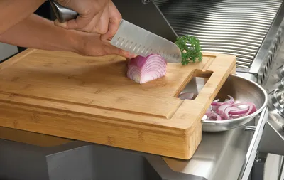 A person is chopping an onion on a wooden cutting board next to a grill.