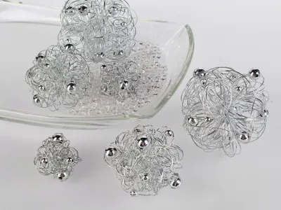 Several decorative wire balls with silver beads are arranged on a clear glass dish filled with small clear beads, with some balls placed outside the dish on a light gray surface.