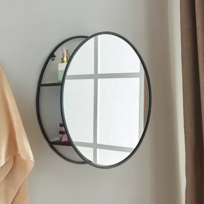 A round black wall mirror with a shelf, mounted on a light gray wall. A beige towel is draped on the left side of the mirror.