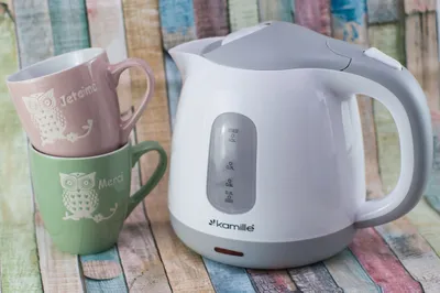 A white and gray electric kettle sits next to two stacked mugs on a colorful wooden background.