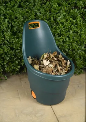 A green garden cart filled with leaves sits on a stone patio in front of a green hedge.