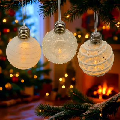 Three illuminated Christmas ornaments hanging from a pine branch, with a blurred background of a Christmas tree and a fireplace.