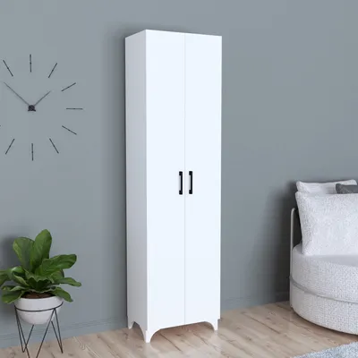A tall, white cabinet with two doors stands against a light gray wall. A plant in a pot is to the left, and a round, textured ottoman is to the right.