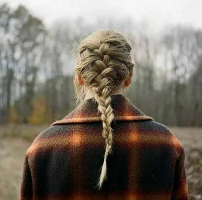 A person from behind wearing a plaid coat and a braided hairstyle, standing in an outdoor setting with trees in the background.