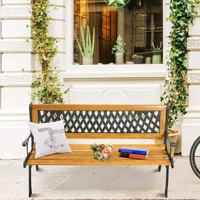 A wooden park bench with a metal frame is placed on a sidewalk in front of a building with ivy growing on the walls. A pillow with a line drawing and a small bouquet of flowers are on the bench.