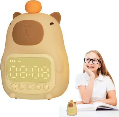 A girl wearing glasses is looking up at a cute capybara-shaped alarm clock. The clock is on a desk with an open book.