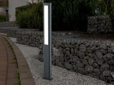 A modern LED bollard light is positioned on a gravel path next to a gabion wall filled with stones.