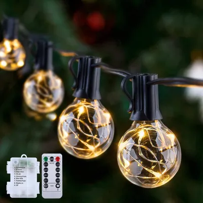 A string of decorative LED lights with clear globe bulbs, shown in a close-up view with a blurred green background resembling a Christmas tree. A battery pack and remote control are visible in the lower left corner.