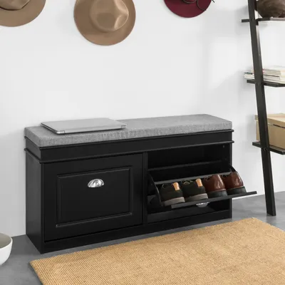 A black shoe bench with a gray cushioned seat, a drawer on the left, and shoe storage compartments on the right. Hats are hanging on the wall behind it, and a ladder shelf is to the right.