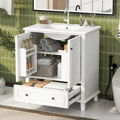 A white bathroom vanity with its doors and drawer open, revealing storage shelves with towels, toiletries, and a basket. The vanity is set against a green tiled wall with a mirror above it.