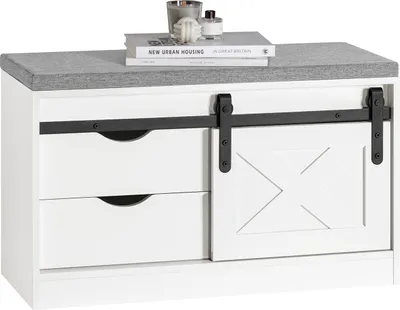 A white shoe bench with a gray upholstered top, featuring two drawers and a sliding barn door with a decorative 'X' pattern. Books and decorative items are placed on top.
