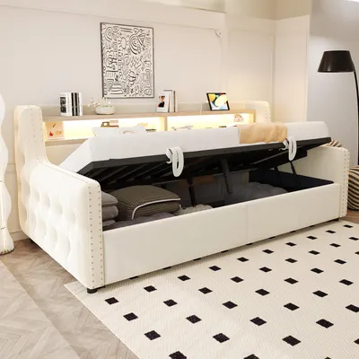 A beige upholstered daybed with its storage compartment open, revealing folded bedding inside. The daybed is placed on a rug with a black diamond pattern in a well-lit room.