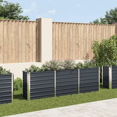A row of dark grey metal raised garden beds filled with plants, set against a wooden fence and a light-colored wall.