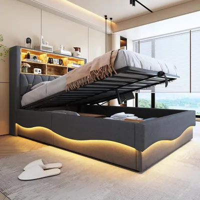 A modern bedroom with a grey upholstered bed frame, featuring a lifted mattress revealing storage space underneath. The bed has integrated LED lighting along the base and a headboard with shelves.