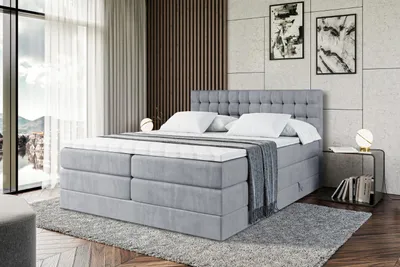 A modern bedroom with a large gray boxspring bed as the focal point, set against a textured wall and large window with sheer curtains.
