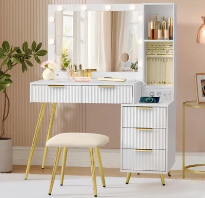 A white vanity table with a lighted mirror, drawers, and a stool, set in a room with a textured wall and a side table.