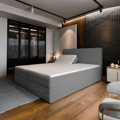 A bedroom scene featuring a large gray boxspring bed with a mattress and a headboard. The bed is positioned in the center of the frame, with a closet visible in the background to the left and a nightstand to the right. The room has wooden flooring and mode