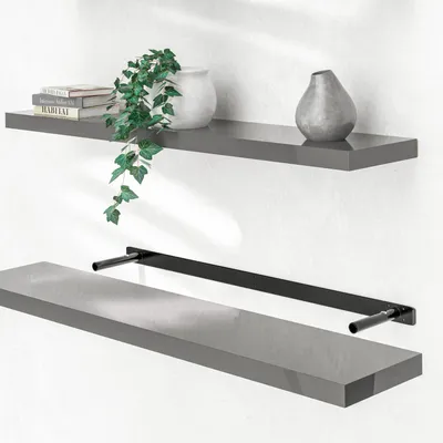 Two gray floating shelves are shown mounted on a white wall. The top shelf has books, a white vase with a plant, and a gray vase. The bottom shelf is shown with its black mounting bracket underneath.