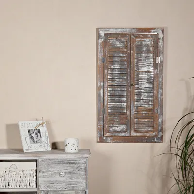 A decorative wooden mirror with shutter-style doors is mounted on a light-colored wall above a distressed wooden dresser. A framed picture and a small decorative pot are on the dresser.