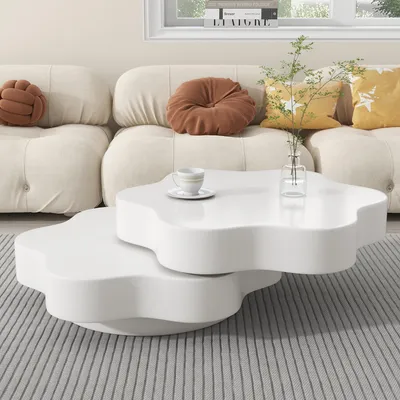A white, flower-shaped coffee table with two tiers is placed on a striped rug in front of a cream-colored sofa. Decorative pillows in brown and yellow are on the sofa, and a small vase with a plant and a cup of coffee are on the table.