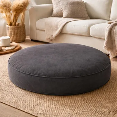 A dark gray round floor cushion is placed on a beige rug in a living room setting, with a white sofa and decorative elements in the background.